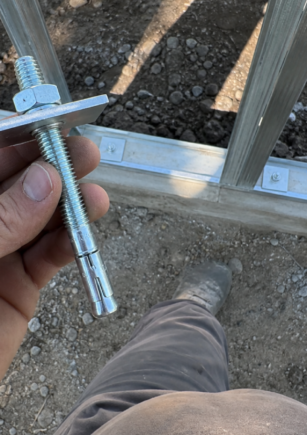 Perno de expansión en uso, sujeto con la mano sobre estructura metálica en obra con grava y zanja