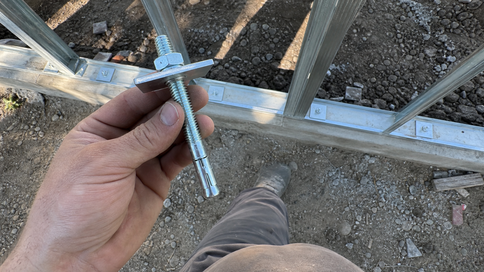 Perno de expansión en uso, sujeto con la mano sobre estructura metálica en obra con grava y zanja